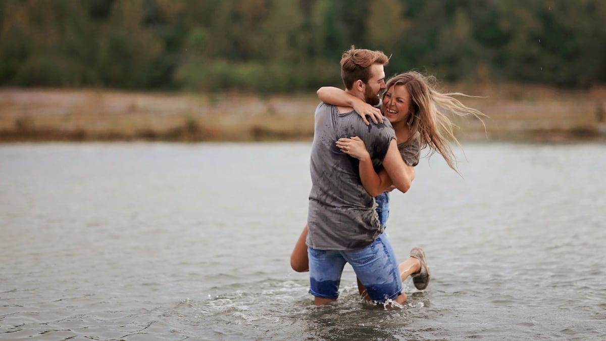 una pareja de un hombre y una mujer, riendo en un lago, mientras él la toma y la eleva.