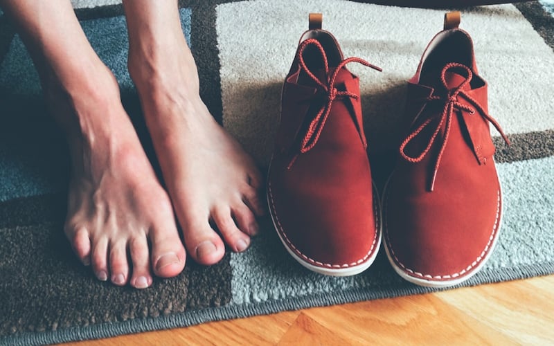 Un par de pies desnudos están posados sobre una alfombra y al lado de unos zapatos de gamuza rojos